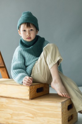 Child in cosy blue knitwear sitting on wooden boxes in a minimal studio — Scandinavian kids fashion editorial. 