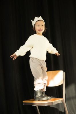 Child standing on a chair under warm studio light, wearing modern kidswear — playful stage-inspired fashion editorial. 