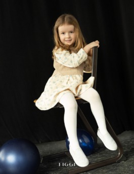 Child standing on a chair under warm studio light, wearing modern kidswear — playful stage-inspired fashion editorial. 