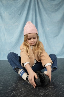 Girl wearing pink knit hat and jeans sitting cross-legged in soft natural light – kids fashion editorial photography by Miriam Dang